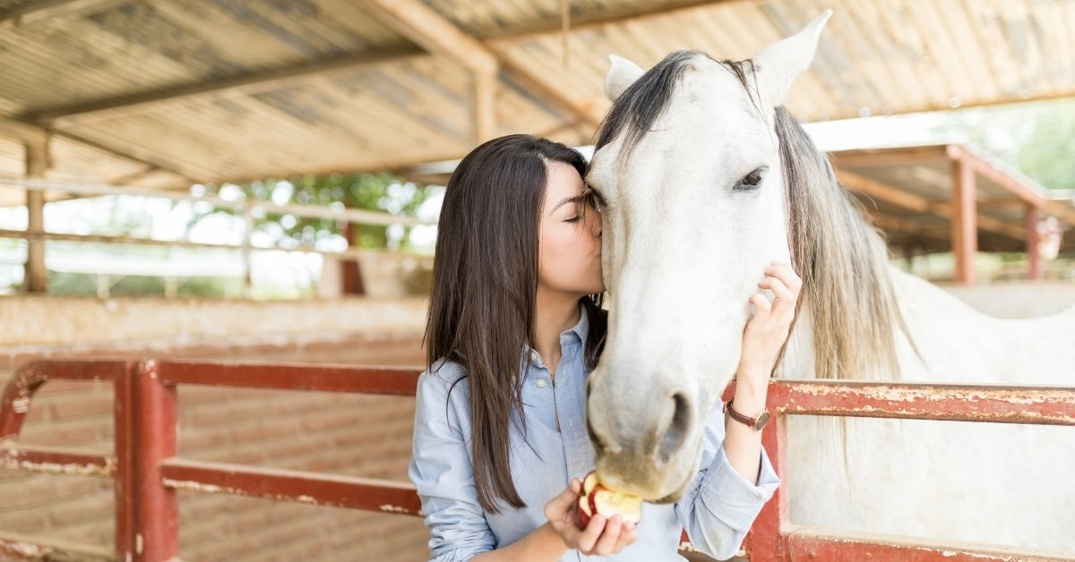 A person kissing a white horse's face while holding an apple near its mouth in a stable.